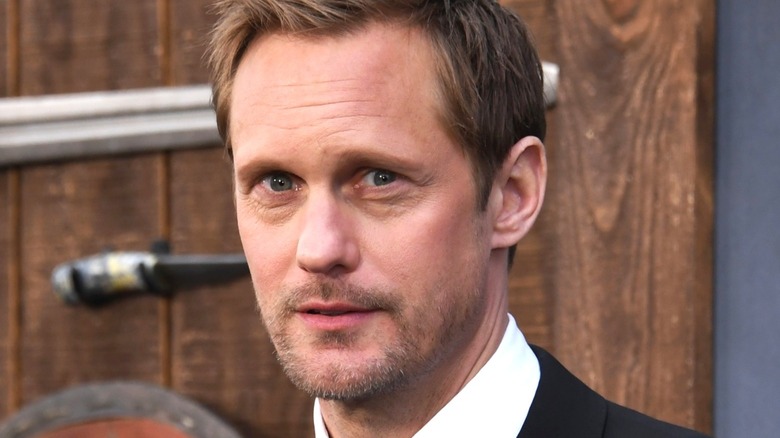 Alexander Skarsgård stands in front of a wooden background