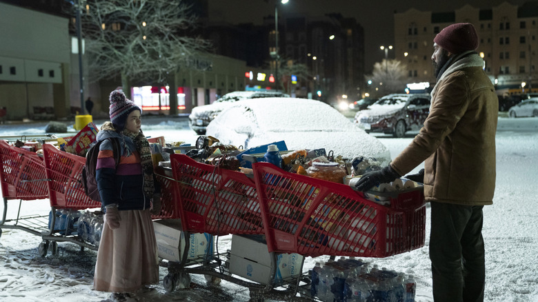 Kirsten talks to Jeevan in the parking lot, standing by a row of shopping carts full of groceries.