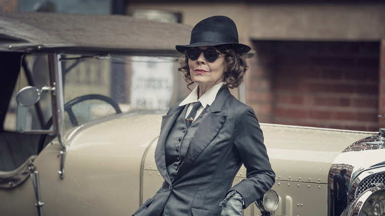 Polly Gray in suit, sunglasses, and hat leaning against hood of tan roadster