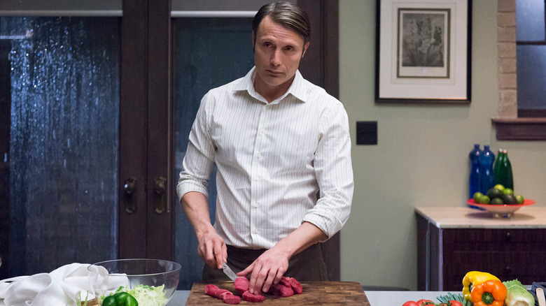 Hannibal slicing red meat at a long kitchen counter surrounded by dinner fixings in Hannibal