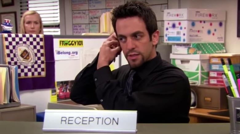 Ryan working at the receptionist's desk while Angela watches him from behind a cubicle wall in The Office