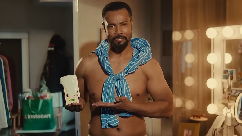 Isaiah Mustafa in a trailer in character as the Old Spice guy, his phone gushing foam, an Instacart tote bag visible behind him