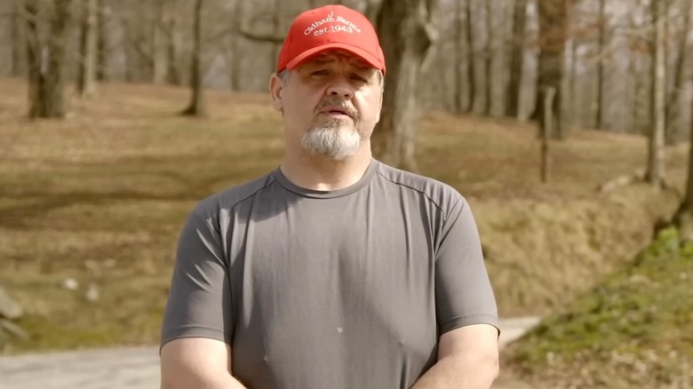 Chris Combs standing in wooded area with red hat and grey shirt