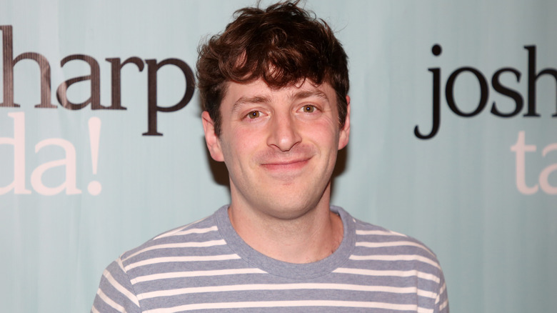 Comedic performer Alex Edelman smiling and posing in front of a light blue wall with black and white letters