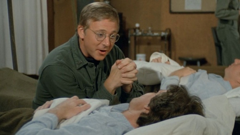 Father Mulcahy sits down while talking to a wounded soldier in M*A*S*H (1982)